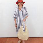 Woman in a striped dress and red hat holding a straw bag against a white wall.