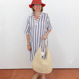 Woman in a striped dress and red hat holding a straw bag against a white wall.