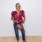 Woman wearing a burgundy floral blouse and blue jeans against a white wall.