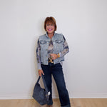 Woman wearing a denim jacket and jeans, holding a gray bag against a white wall.