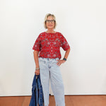 Woman wearing a red patterned blouse and light blue jeans, holding a denim jacket against a white wall.