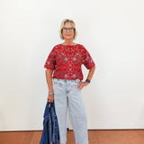Woman wearing a red patterned blouse and light blue jeans, holding a denim jacket against a white wall.