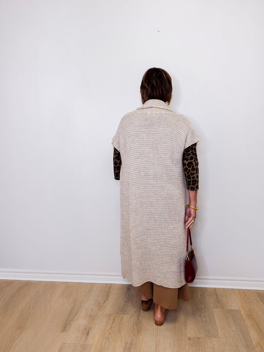 Person wearing a long beige cardigan with leopard print sleeves against a white wall.
