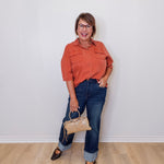 Woman wearing an orange shirt and blue jeans, holding a beige handbag against a white wall.