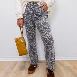 Person wearing leopard print pants and holding a brown bag against a white wall.