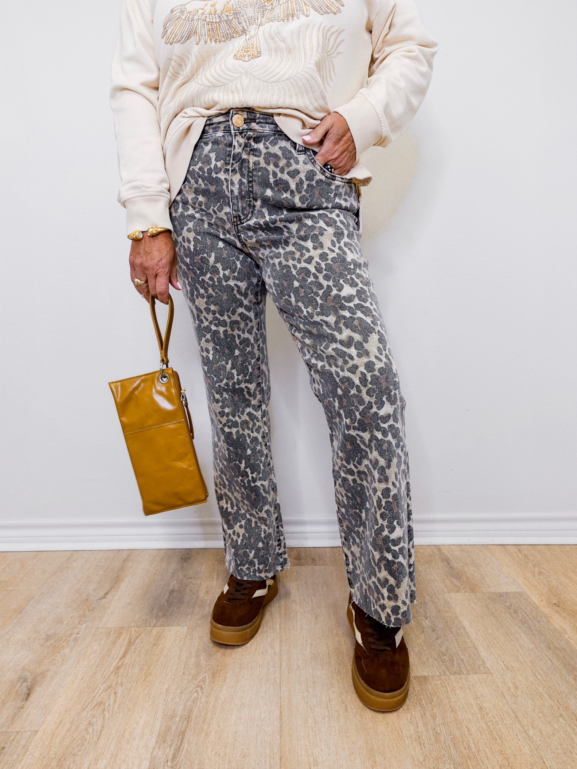 Person wearing leopard print pants and holding a brown bag against a white wall.