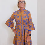 Woman wearing a brown dress with blue patterns against a white wall