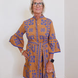 Woman wearing a brown dress with blue patterns against a white wall