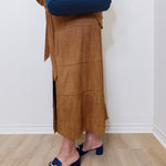 Person wearing a brown suede skirt and blue shoes, holding a blue bag against a white wall.