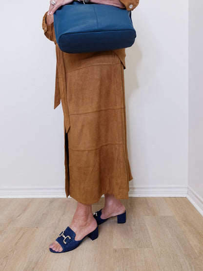 Person wearing a brown suede skirt and blue shoes, holding a blue bag against a white wall.