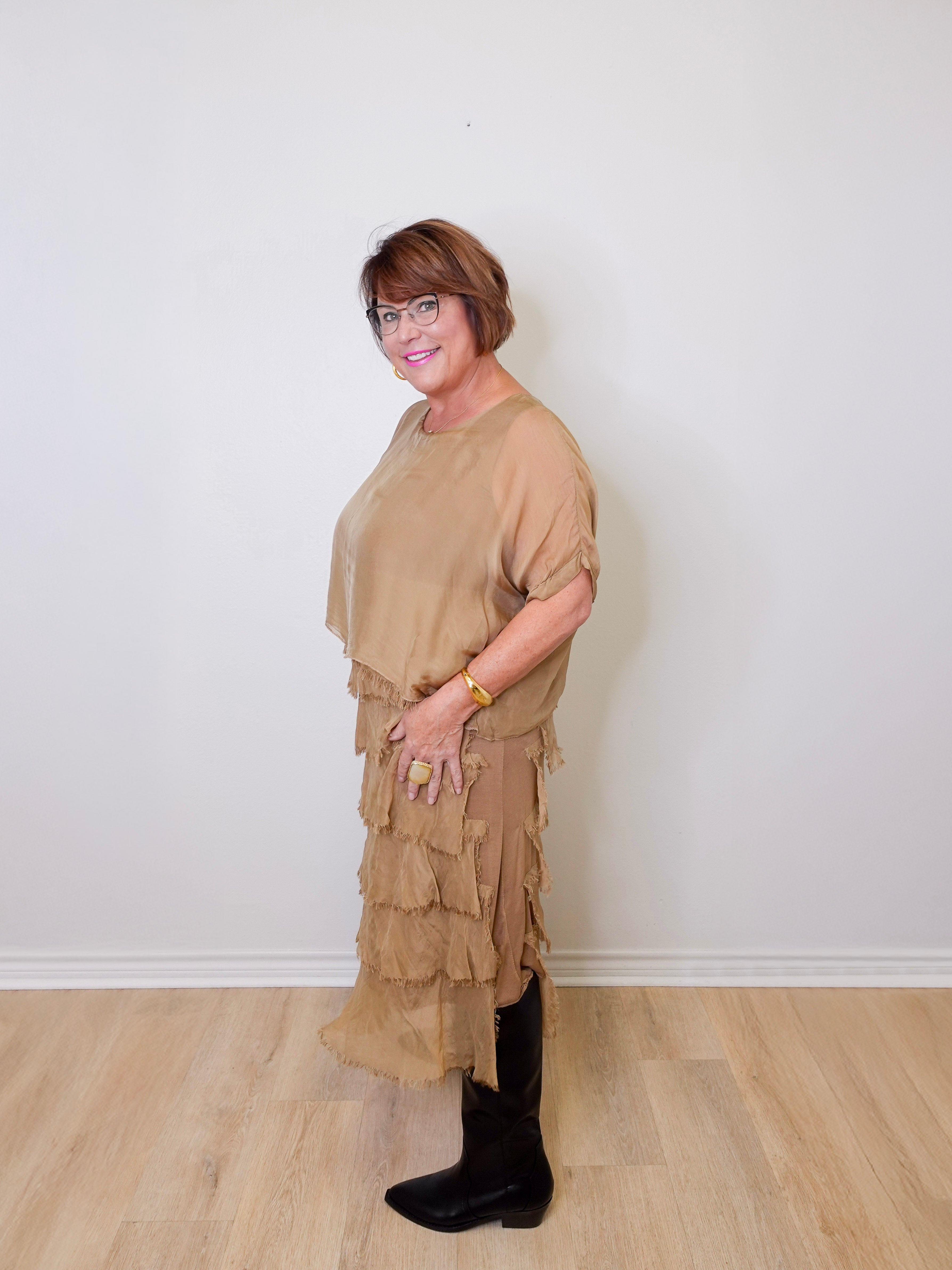 Woman wearing a beige layered dress and black boots against a white wall.