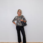 Woman in floral blouse and black pants holding a black bag against a white wall