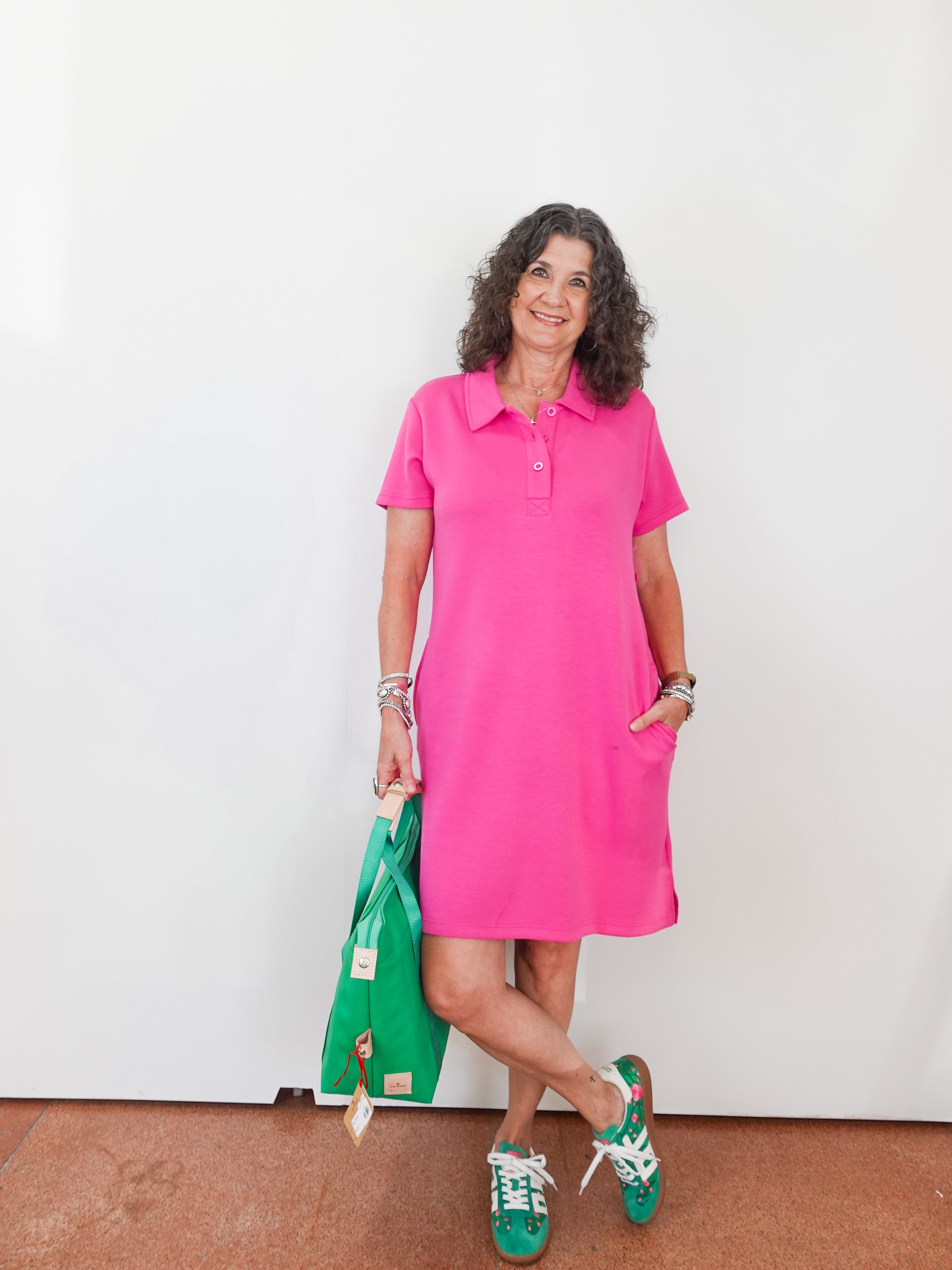 Woman wearing a pink dress holding a green bag against a white wall.