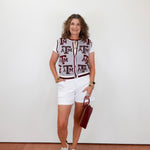 Woman wearing a patterned top and white shorts standing against a plain background