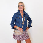 Woman wearing a denim jacket and floral skirt holding a clear handbag against a white wall.