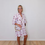 Woman wearing a white dress with red floral pattern against a plain white wall.