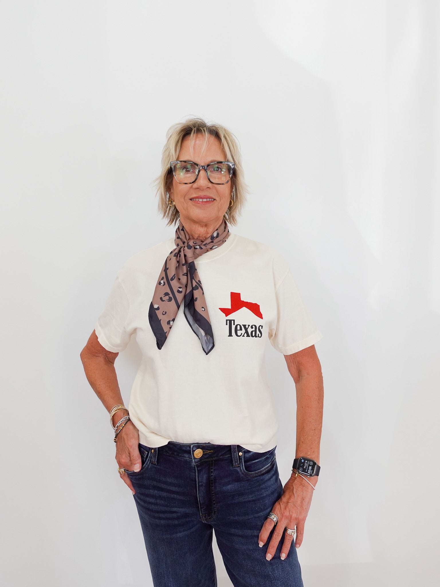Person wearing a white t-shirt with a red and black logo, blue jeans, and a patterned scarf.