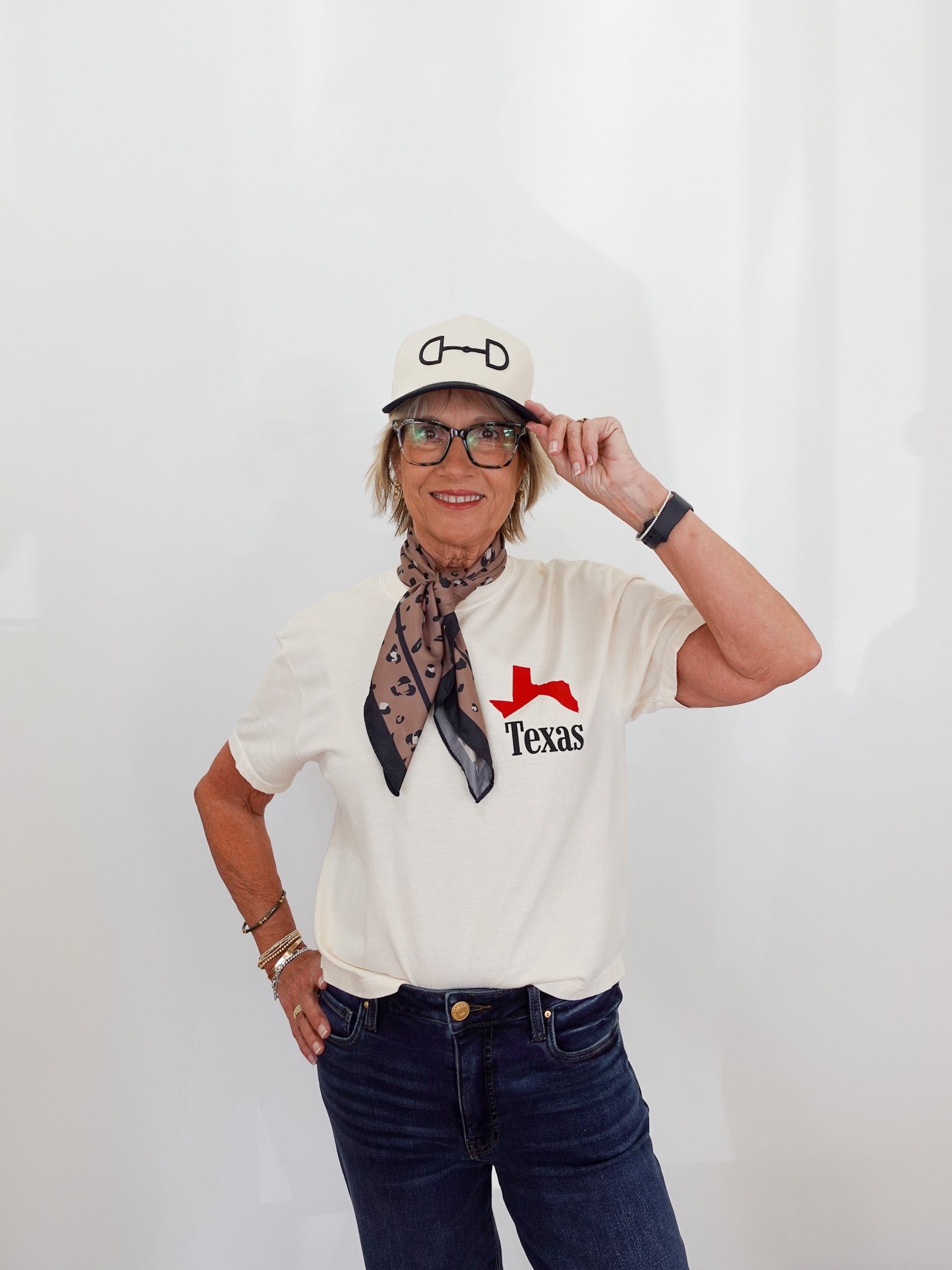 Person wearing a white 'Texas' t-shirt, dark jeans, and a cap with a scarf around their neck against a white background.