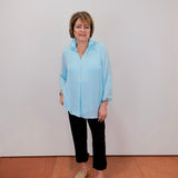 Woman wearing a light blue shirt and black pants standing against a white wall.