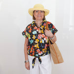 Woman wearing a colorful floral blouse, white pants, and a yellow hat, holding a straw bag against a white background.