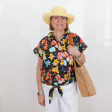 Woman wearing a colorful floral blouse, white pants, and a yellow hat, holding a straw bag against a white background.
