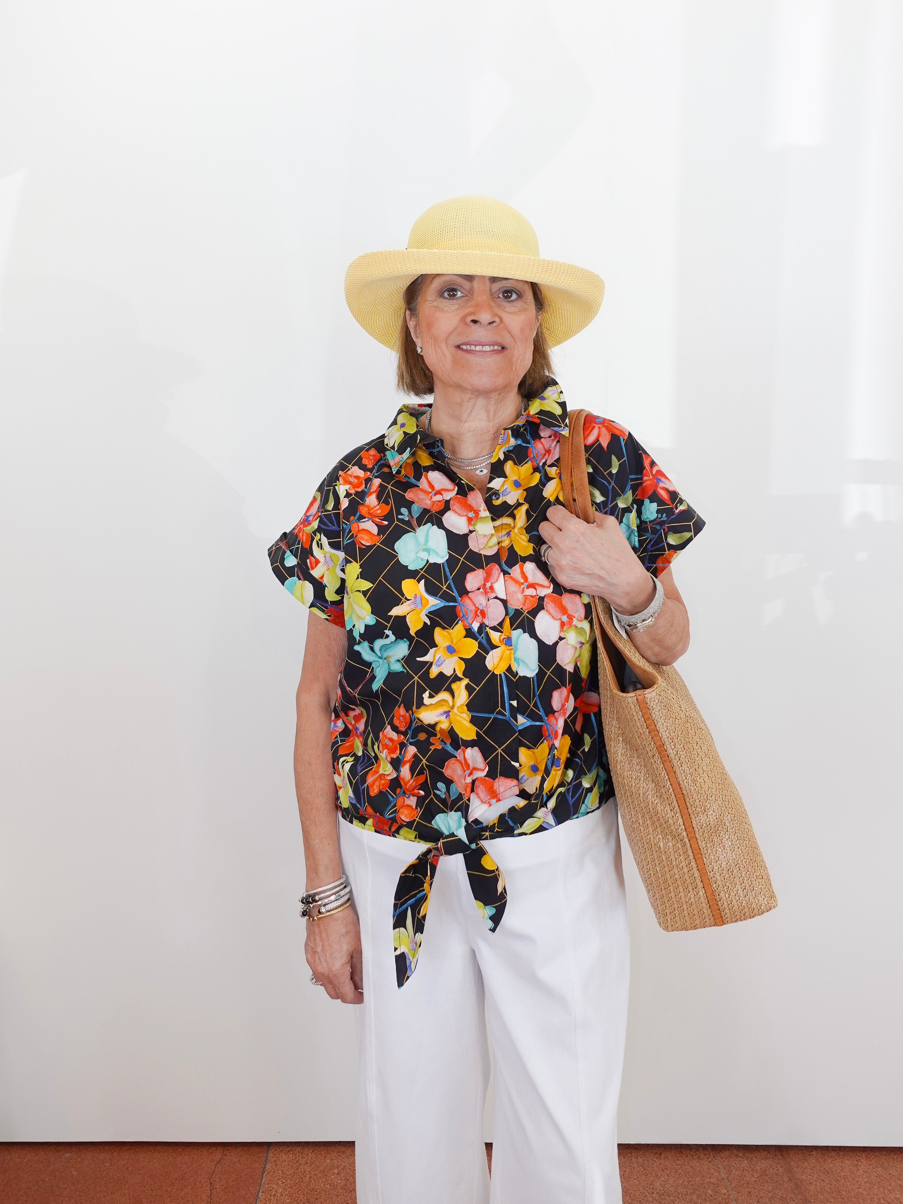 Woman wearing a colorful floral blouse, white pants, and a yellow hat, holding a straw bag against a white background.