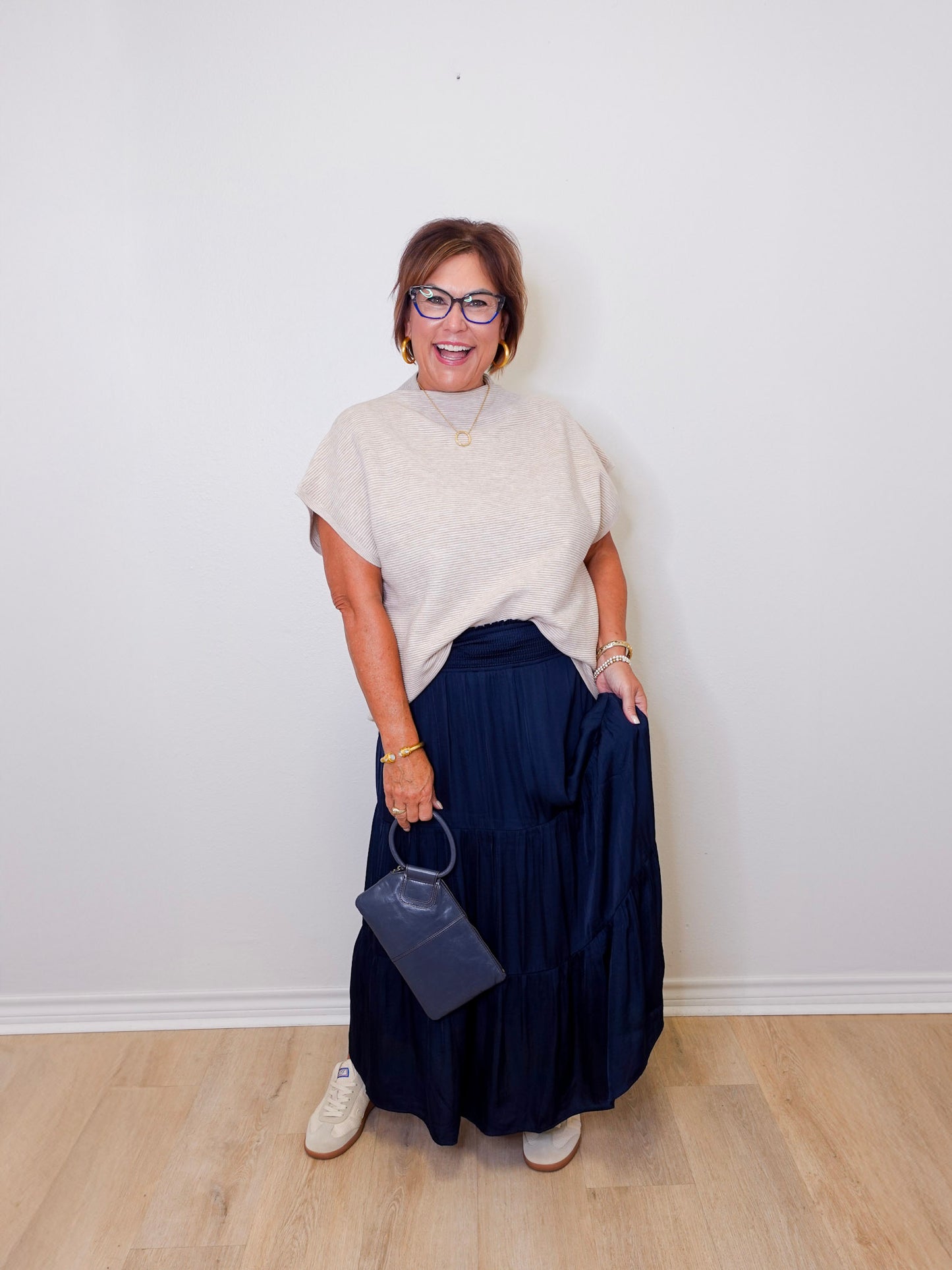 Woman wearing a beige sweater and navy skirt holding a blue handbag against a white wall.