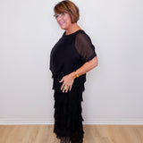 Woman wearing a black dress with sheer sleeves against a white wall.