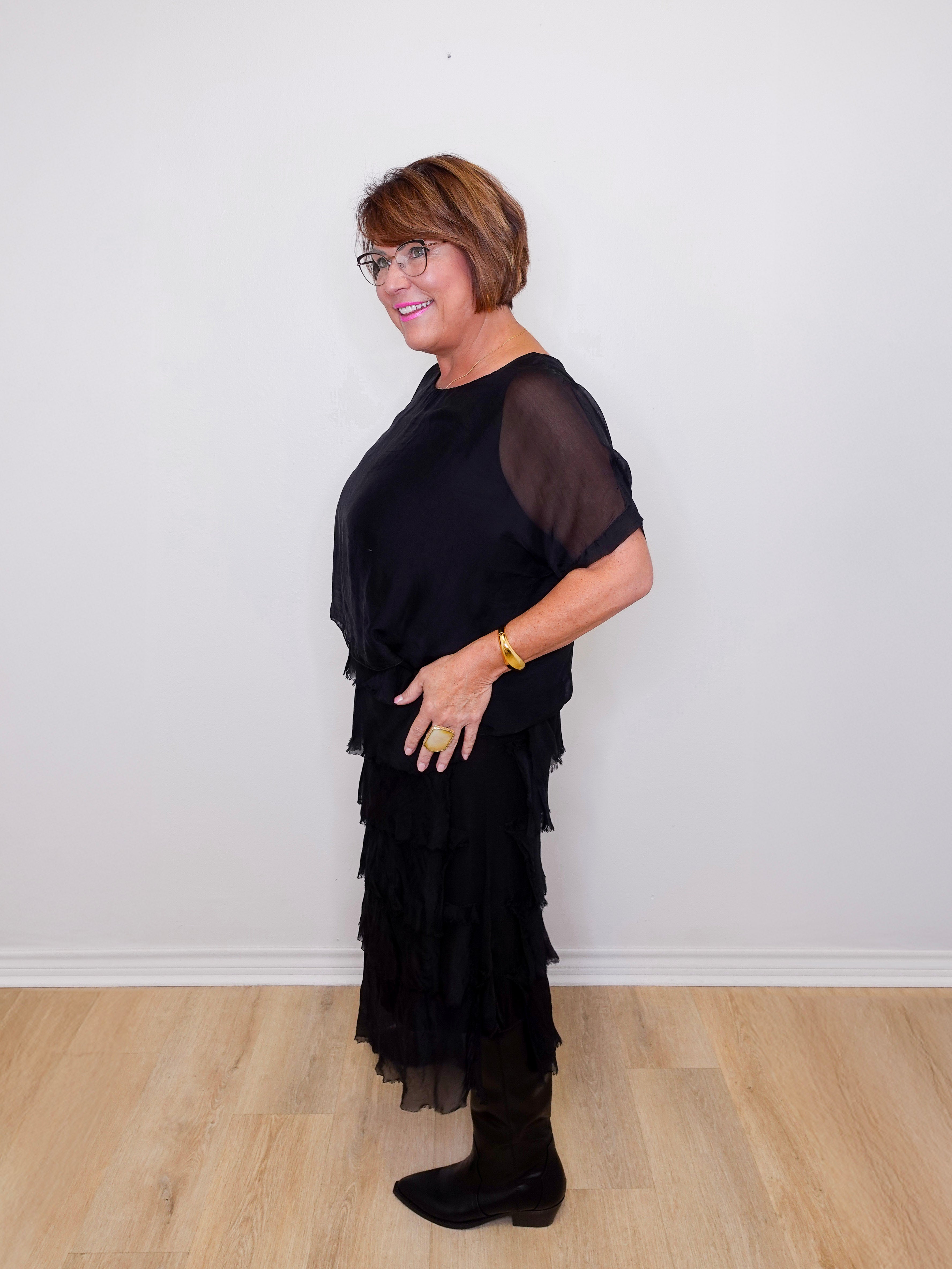 Woman wearing a black dress with sheer sleeves against a white wall.