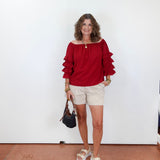 Woman wearing a red blouse with ruffled sleeves, beige shorts, and sandals, holding a black handbag against a white wall.