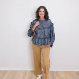 Woman wearing a blue patterned top and beige pants standing against a white wall.