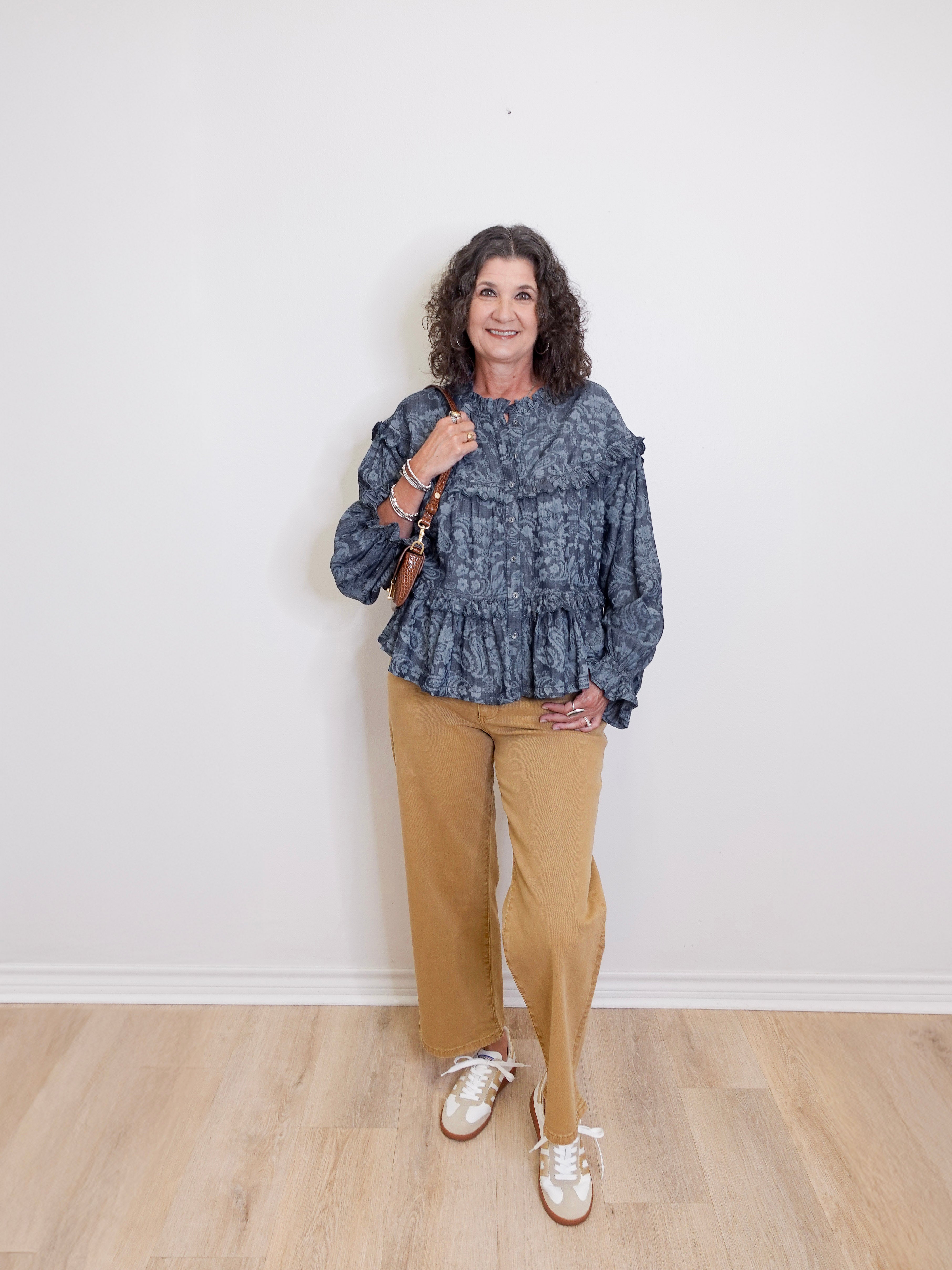 Woman wearing a blue patterned top and beige pants standing against a white wall.
