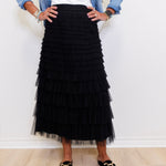 Person wearing a black tiered skirt with a white top and denim jacket against a white wall.