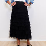 Person wearing a black tiered skirt with a white top and denim jacket against a white wall.