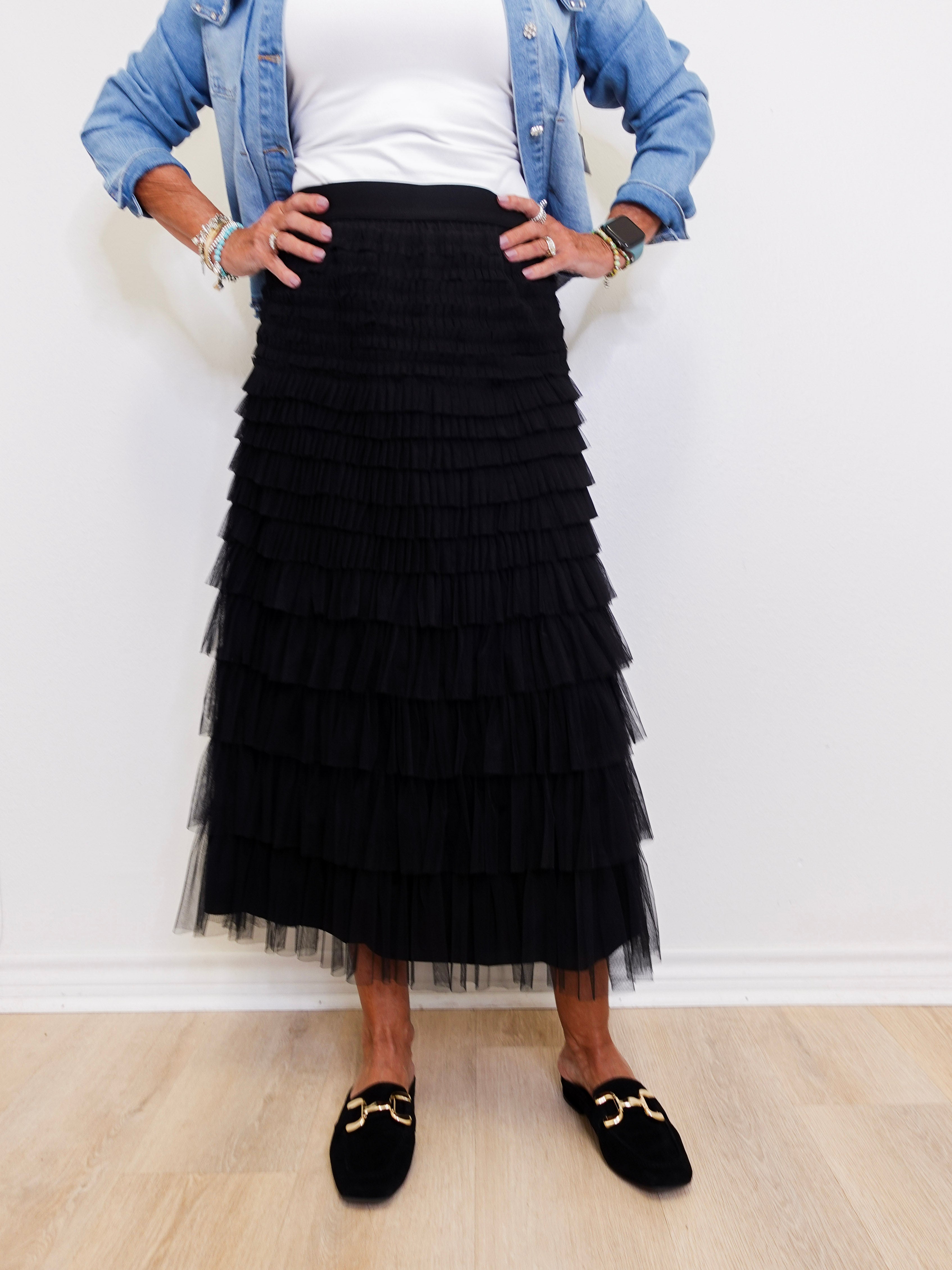 Person wearing a black tiered skirt with a white top and denim jacket against a white wall.