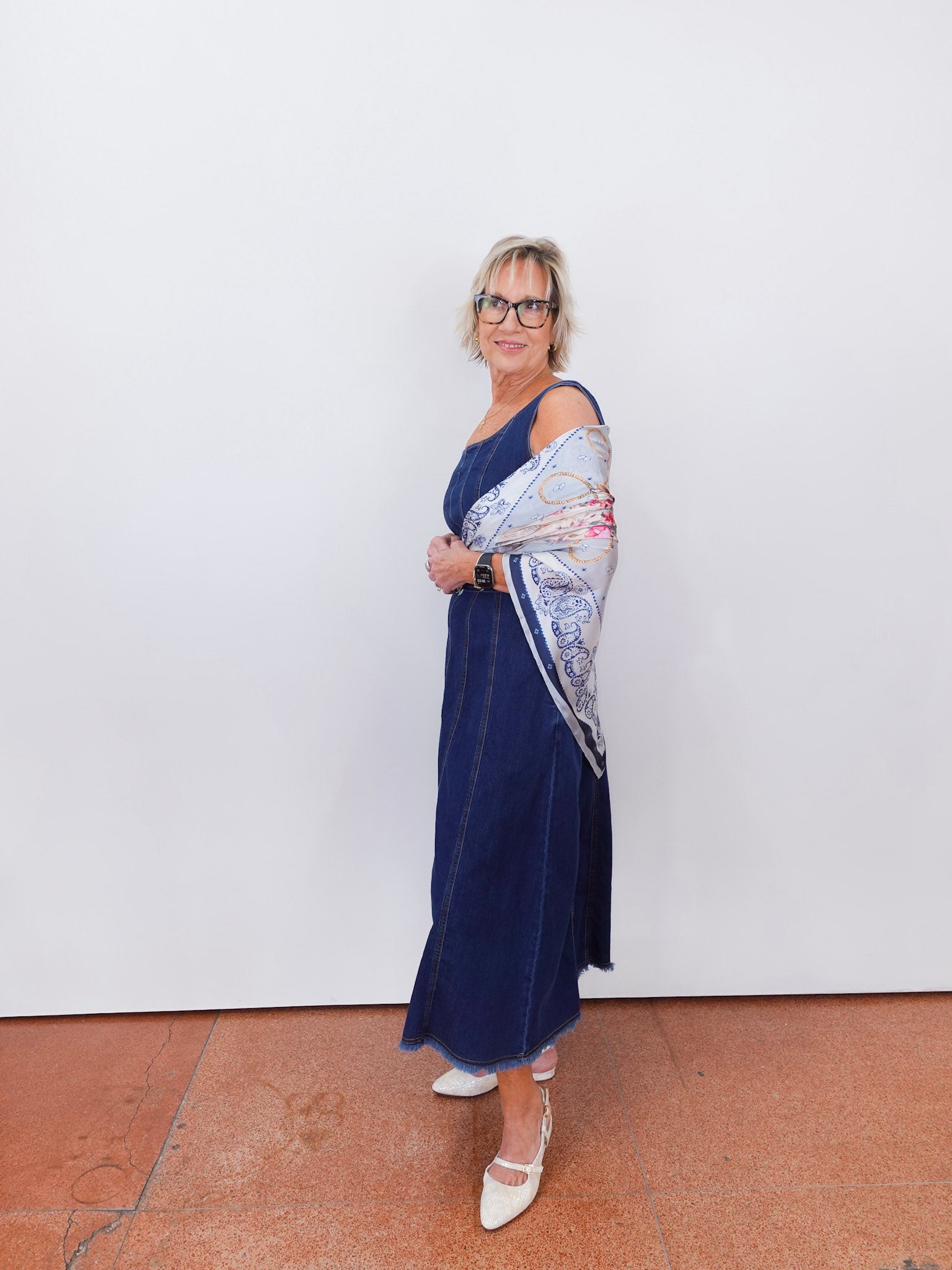 Woman wearing a blue dress with a patterned shawl against a white wall.