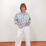 Woman wearing a floral blouse and white pants against a plain background