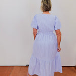 Woman wearing a light blue striped dress standing against a white wall.