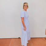Woman wearing a light blue dress with white sneakers against a white wall.