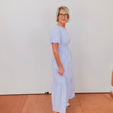 Woman wearing a light blue dress with white sneakers against a white wall.
