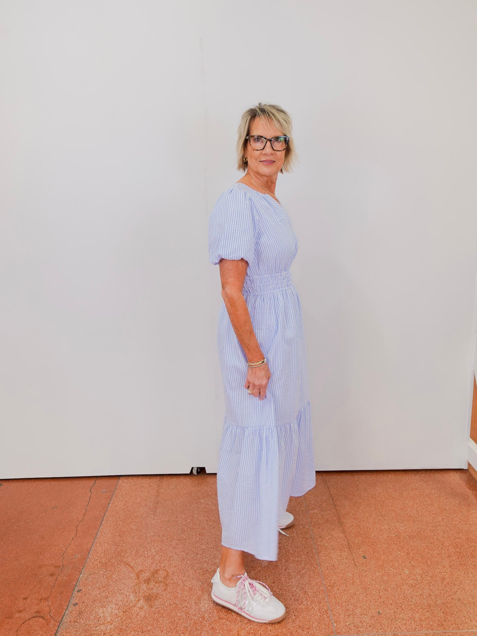 Woman wearing a light blue dress with white sneakers against a white wall.