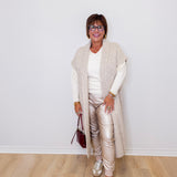 Woman wearing a long beige cardigan, white top, and gold pants against a white wall.