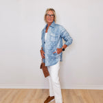 Person wearing a blue denim jacket, white pants, and brown shoes against a white wall.