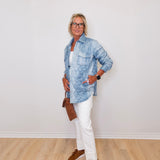 Person wearing a blue denim jacket, white pants, and brown shoes against a white wall.