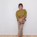 Woman wearing a green patterned sweater and beige pants against a white wall.