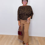 Woman wearing a leopard print top and beige pants, holding a red handbag against a white wall.