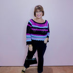 Woman wearing a colorful sweater and black pants against a plain background