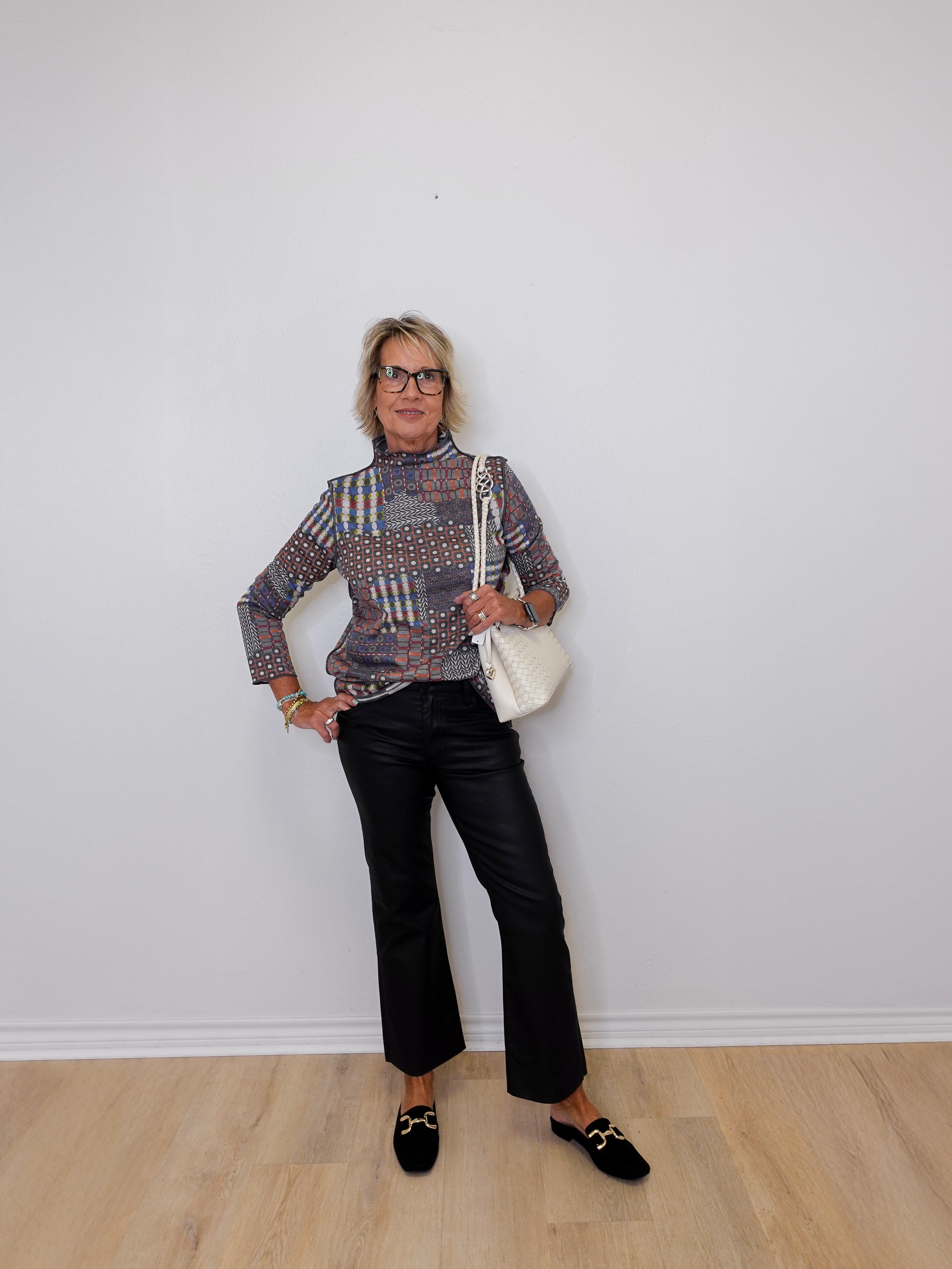 Person wearing a patterned blouse, black pants, and black shoes against a white wall.