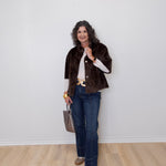 Woman wearing a brown jacket, white shirt, blue jeans, and gold shoes against a white wall.