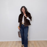 Woman wearing a brown jacket, white shirt, blue jeans, and gold shoes against a white wall.